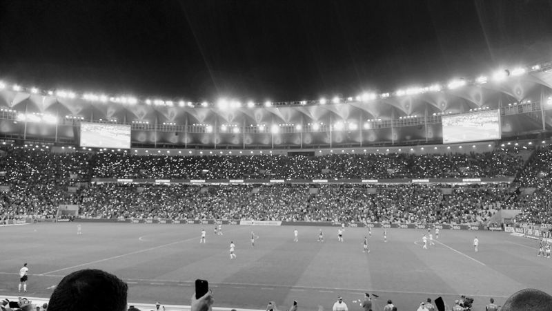 Maracanã lotado à noite durante jogo do Flamengo, com jogadores em campo ao longe, em preto e branco